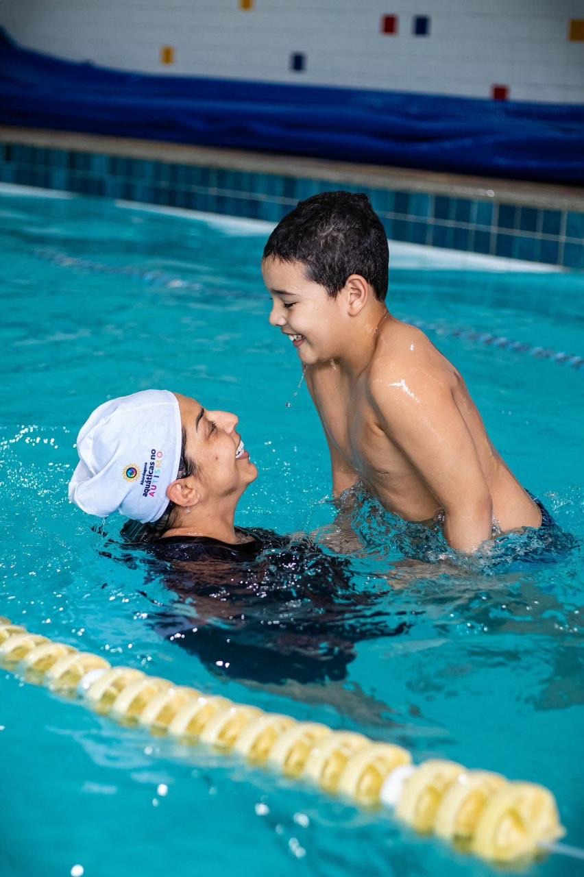 Instrutora de touca branca sorri para um menino que ela sustenta na água da piscina